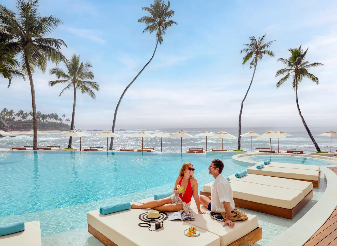 A couple relaxing on a poolside daybed beside an infinity pool, with palm trees and the Indian Ocean behind them